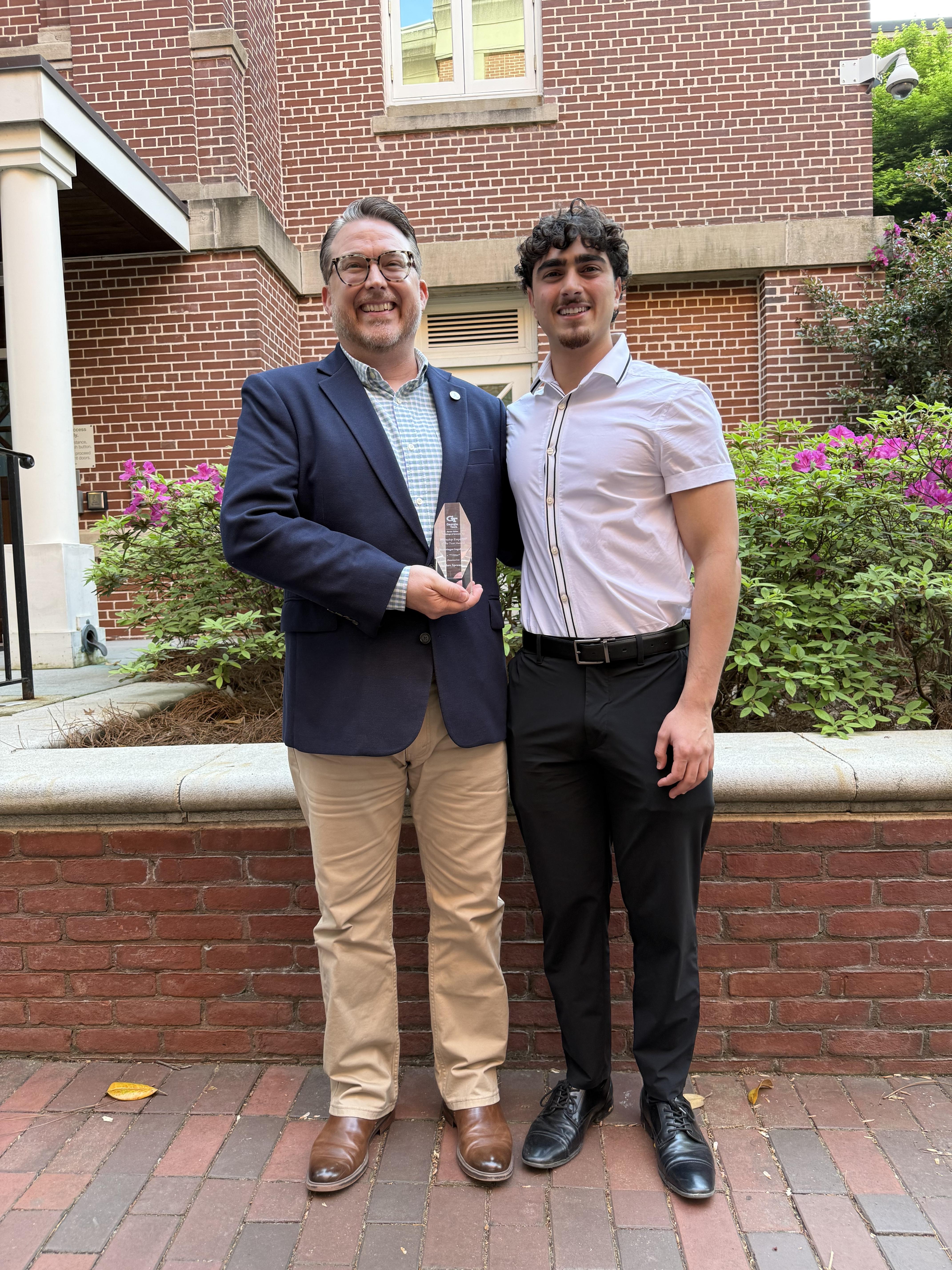 A man and a male college student shake hands and hold up an award.