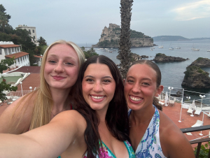 Three girls on a clifftop overlooking blue water taking a selfie.