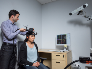 Researcher adjusting a device on another person’s head in a lab, with a computer displaying brain imaging data and a mounted camera in the background.