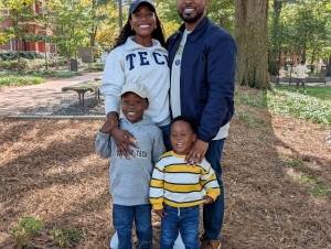A family with two young boys stands in front of a tree.