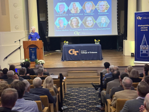 a woman at a podium in front of a screen highlighting all of the Distinguished Alumni Award winners.