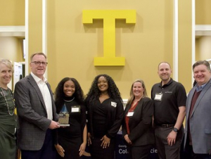 Seven people pose with an award in front of a giant letter T.