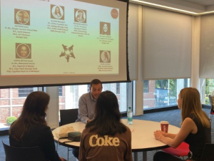 A man sitting in front of a screen talks with 3 students at a round table.