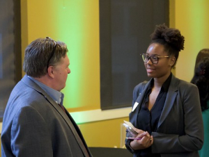 A man and young woman talk in front of two windows.